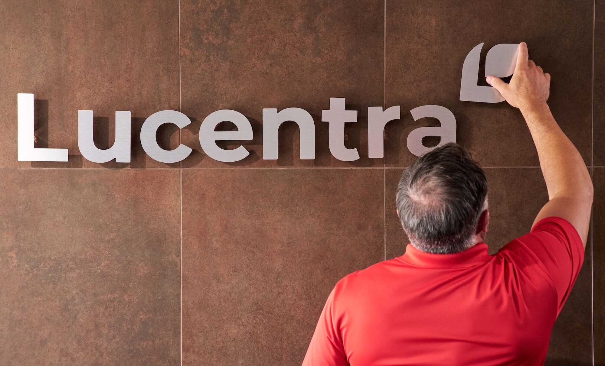 Man in red shirt adjusts sign reading "Lucentra" on wall